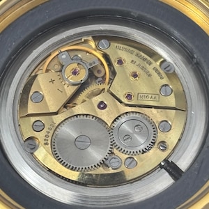 K&ouml;nnte beinhalten: Nahaufnahme eines Uhrwerks mit goldfarbenen Zahnr&auml;dern, R&auml;dern und Juwelen. Das Innenleben der Uhr ist sichtbar, einschlie&szlig;lich des Textes ULYSSE NARDIN UNION und der Anzahl der Juwelen. Die Uhr hat eine goldfarbene L&uuml;nette.
