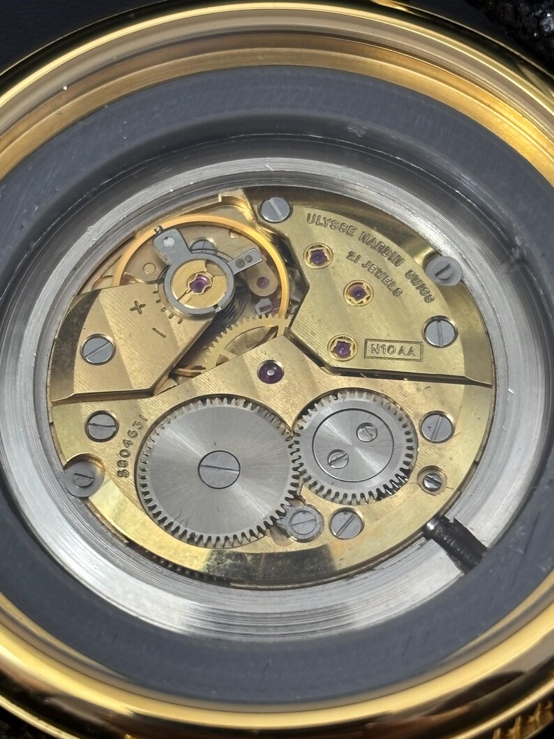 K&ouml;nnte beinhalten: Nahaufnahme eines Uhrwerks mit goldfarbenen Zahnr&auml;dern, R&auml;dern und Juwelen. Das Innenleben der Uhr ist sichtbar, einschlie&szlig;lich des Textes ULYSSE NARDIN UNION und der Anzahl der Juwelen. Die Uhr hat eine goldfarbene L&uuml;nette.