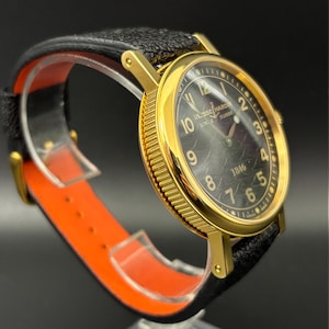 K&ouml;nnte beinhalten: Eine goldfarbene Armbanduhr mit schwarzem Zifferblatt und schwarzem Lederarmband. Das Zifferblatt zeigt den Markennamen "ULYSSE NARDIN LOCLE SUISSE" und das Jahr "1846". Die Uhr wird auf einem orangefarbenen und transparenten St&auml;nder pr&auml;sentiert.