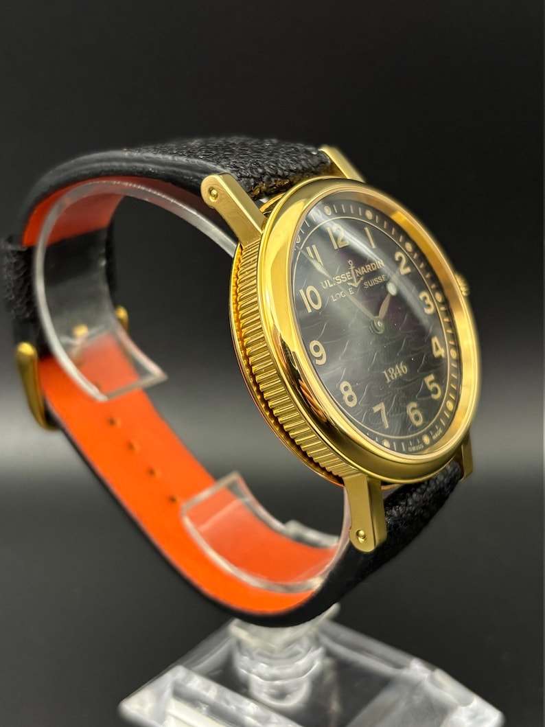 K&ouml;nnte beinhalten: Eine goldfarbene Armbanduhr mit schwarzem Zifferblatt und schwarzem Lederarmband. Das Zifferblatt zeigt den Markennamen "ULYSSE NARDIN LOCLE SUISSE" und das Jahr "1846". Die Uhr wird auf einem orangefarbenen und transparenten St&auml;nder pr&auml;sentiert.