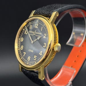 K&ouml;nnte beinhalten: Eine goldfarbene Ulysse Nardin-Uhr mit schwarzem Zifferblatt und schwarzem Lederarmband. Die Uhr hat goldene Ziffern und Zeiger, mit dem Markennamen und "SUISSE" sichtbar. Die Uhr wird auf einem transparenten St&auml;nder mit orangefarbenem Innenraum pr&auml;sentiert.