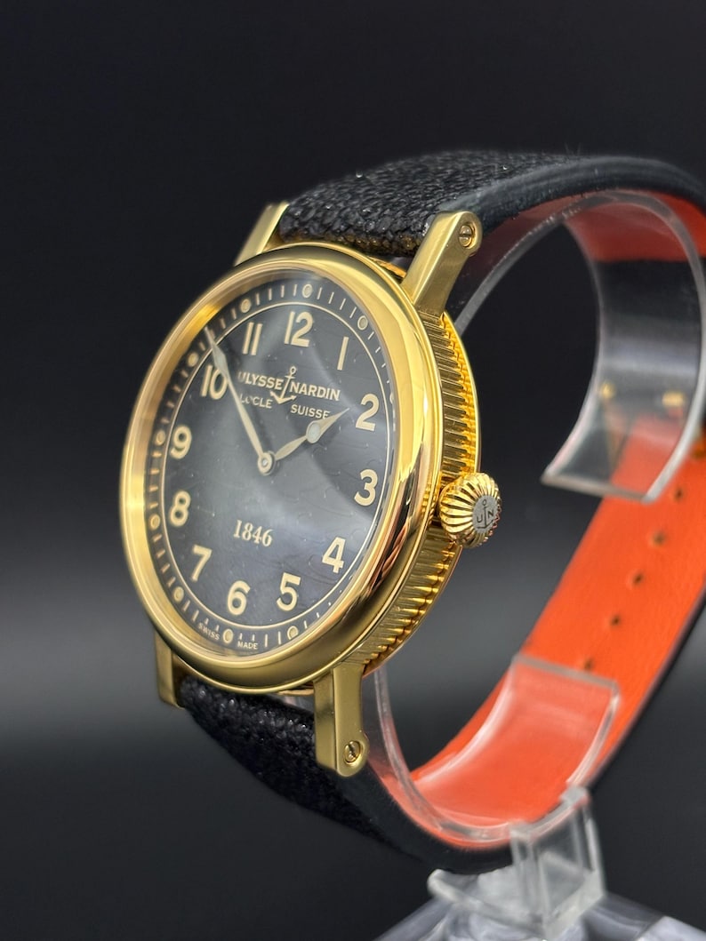 K&ouml;nnte beinhalten: Eine goldfarbene Ulysse Nardin-Uhr mit schwarzem Zifferblatt und schwarzem Lederarmband. Die Uhr hat goldene Ziffern und Zeiger, mit dem Markennamen und "SUISSE" sichtbar. Die Uhr wird auf einem transparenten St&auml;nder mit orangefarbenem Innenraum pr&auml;sentiert.