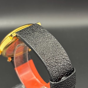 K&ouml;nnte beinhalten: Nahaufnahme einer goldfarbenen Armbanduhr mit einem schwarzen, strukturierten Lederarmband. Das Zifferblatt ist teilweise sichtbar, und das Armband hat eine detaillierte, genarbte Oberfl&auml;che. Die Uhr ist vor einem dunklen Hintergrund.