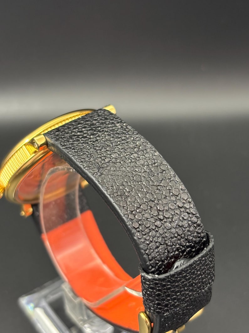 K&ouml;nnte beinhalten: Nahaufnahme einer goldfarbenen Armbanduhr mit einem schwarzen, strukturierten Lederarmband. Das Zifferblatt ist teilweise sichtbar, und das Armband hat eine detaillierte, genarbte Oberfl&auml;che. Die Uhr ist vor einem dunklen Hintergrund.
