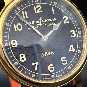 K&ouml;nnte beinhalten: Nahaufnahme einer goldfarbenen Ulysse Nardin Uhr mit schwarzem Zifferblatt. Die Uhr hat goldene Zeiger und Ziffern, mit dem Markennamen und "SUISSE" sichtbar. Das Armband ist aus schwarzem Leder.