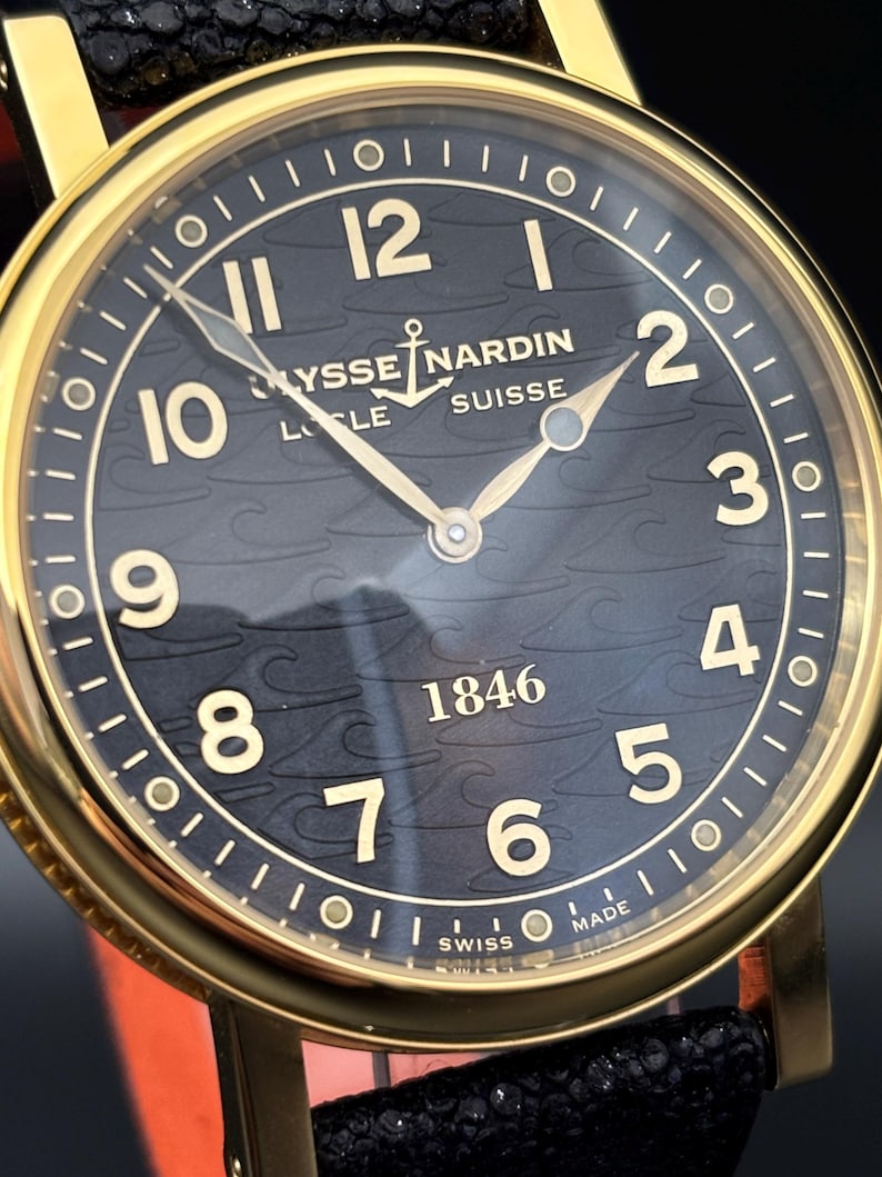 K&ouml;nnte beinhalten: Nahaufnahme einer goldfarbenen Ulysse Nardin Uhr mit schwarzem Zifferblatt. Die Uhr hat goldene Zeiger und Ziffern, mit dem Markennamen und "SUISSE" sichtbar. Das Armband ist aus schwarzem Leder.