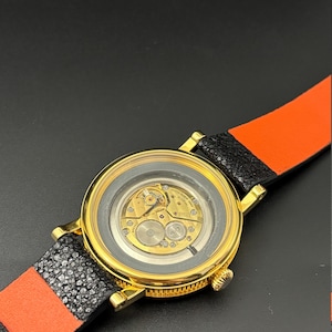 K&ouml;nnte beinhalten: Eine goldfarbene Armbanduhr mit transparentem R&uuml;ckteil, das das Uhrwerk zeigt. Das Armband ist aus schwarzem und orangefarbenem Leder. Das runde Zifferblatt hat eine goldene L&uuml;nette.