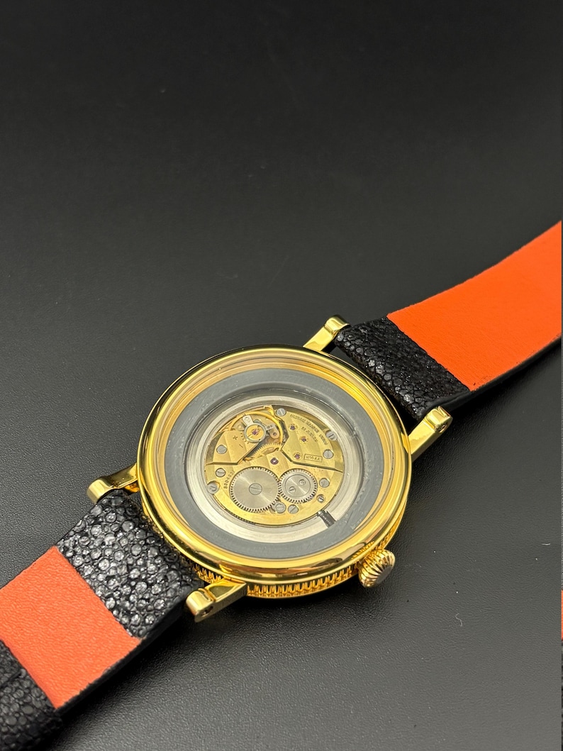 K&ouml;nnte beinhalten: Eine goldfarbene Armbanduhr mit transparentem R&uuml;ckteil, das das Uhrwerk zeigt. Das Armband ist aus schwarzem und orangefarbenem Leder. Das runde Zifferblatt hat eine goldene L&uuml;nette.