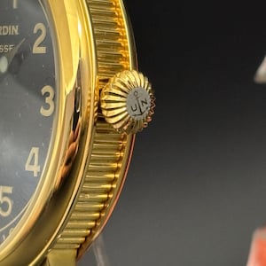 K&ouml;nnte beinhalten: Nahaufnahme einer goldfarbenen Armbanduhr mit schwarzem Zifferblatt. Die Uhr hat gro&szlig;e, goldfarbene Ziffern und eine strukturierte L&uuml;nette. Die Krone ist ebenfalls goldfarben und hat ein Logo mit den Buchstaben "UN" und einem Anker.