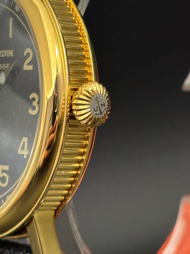K&ouml;nnte beinhalten: Nahaufnahme einer goldfarbenen Armbanduhr mit schwarzem Zifferblatt. Die Uhr hat gro&szlig;e, goldfarbene Ziffern und eine strukturierte L&uuml;nette. Die Krone ist ebenfalls goldfarben und hat ein Logo mit den Buchstaben "UN" und einem Anker.