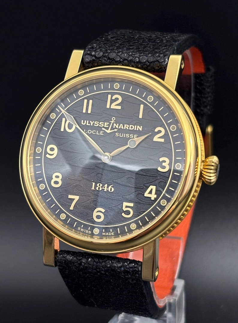 K&ouml;nnte beinhalten: Eine goldfarbene Ulysse Nardin Uhr mit schwarzem Zifferblatt und schwarzem Lederarmband. Das Zifferblatt zeigt goldene Ziffern und Zeiger mit dem Text "ULYSSE NARDIN LOCLE SUISSE 1846". Die Uhr hat ein strukturiertes schwarzes Zifferblatt.