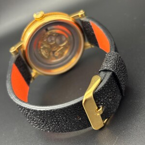 K&ouml;nnte beinhalten: Eine goldfarbene Armbanduhr mit einem strukturierten schwarzen Armband und einer goldenen Schnalle. Das Zifferblatt der Uhr ist sichtbar und zeigt das Innenleben. Das Armband hat ein rot-schwarzes Streifendetail. Die Uhr wird von hinten betrachtet.