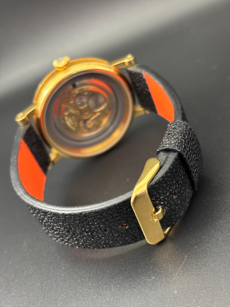 K&ouml;nnte beinhalten: Eine goldfarbene Armbanduhr mit einem strukturierten schwarzen Armband und einer goldenen Schnalle. Das Zifferblatt der Uhr ist sichtbar und zeigt das Innenleben. Das Armband hat ein rot-schwarzes Streifendetail. Die Uhr wird von hinten betrachtet.