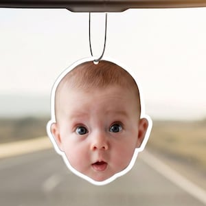May include: A custom air freshener hanging from a rearview mirror, featuring a high-resolution photographic print of a baby's face with wide eyes and a small mouth, cut into the shape of the head with a white border and a silver hanging string.