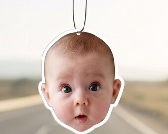 May include: A custom air freshener hanging from a rearview mirror, featuring a high-resolution photographic print of a baby's face with wide eyes and a small mouth, cut into the shape of the head with a white border and a silver hanging string.