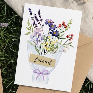 May include: Greeting card featuring a watercolor bouquet of flowers in shades of purple, blue, red, and yellow. The bouquet is wrapped in translucent paper and tied with a purple ribbon. The word "friend" is printed on a tan banner.