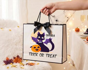 May include: A white rectangular gift bag with black trim and a black ribbon bow. The front features a graphic of a black cat with purple bat wings sitting next to an orange jack-o'-lantern. The text reads SAMARA and TRICK OR TREAT.