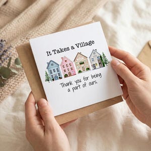 Puede incluir: Una tarjeta de felicitación con el texto "It Takes a Village" y "Thank you for being a part of ours." La tarjeta presenta ilustraciones de casas coloridas. La tarjeta se sostiene en un sobre marrón.