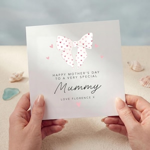 May include: A Mother's Day card held by hands, featuring a white bow with pink heart details and the words "Happy Mother's Day to a very special Mummy" in black script. The card is signed "Love Florence X".