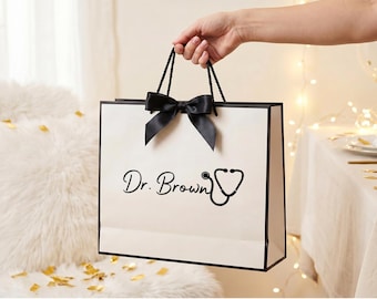 May include: A rectangular white gift bag with black trim and black rope handles. A black satin ribbon bow is attached to the top center. The front features the black text Dr. Brown followed by a black graphic illustration of a stethoscope.