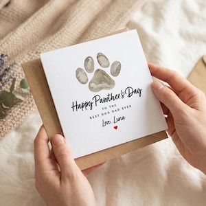 May include: A greeting card with a paw print design and the text "Happy Pawther's Day to the Best Dog Dad Ever, Love, Luna." The card is held in front of a brown envelope and a sprig of lavender.