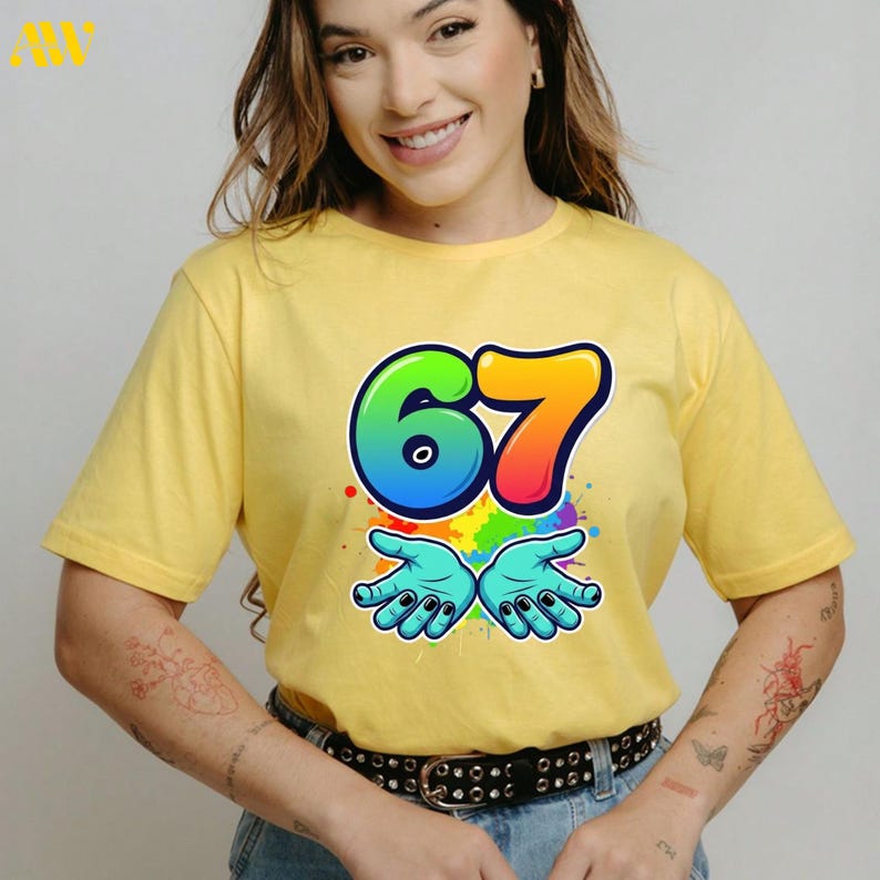 May include: Yellow t-shirt with a colorful graphic of the number 67, outlined in black, with a rainbow gradient. Below the number are two blue hands with black fingernails, and colorful paint splatters.