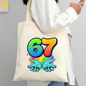 May include: Cream-colored tote bag featuring a colorful graphic of the number 67 in rainbow hues, with open hands below. The design includes paint splatters and black nail polish on the hands.