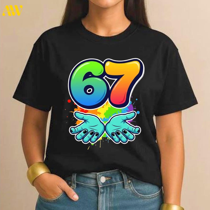 May include: Black t-shirt featuring a colorful graphic design. The design includes the number "67" in rainbow colors, outlined in white, with two cartoon hands below. The hands are light blue with black fingernails, set against a colorful splatter background.