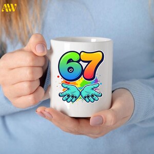May include: White ceramic mug featuring the number "67" in a colorful, gradient design. Below the number are two turquoise hands with black fingernails, set against a backdrop of colorful paint splatters. The mug is held by a person.
