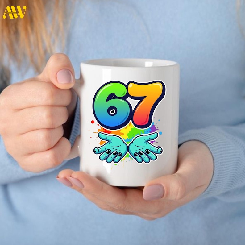 May include: White ceramic mug featuring the number "67" in a colorful, gradient design. Below the number are two turquoise hands with black fingernails, set against a backdrop of colorful paint splatters. The mug is held by a person.