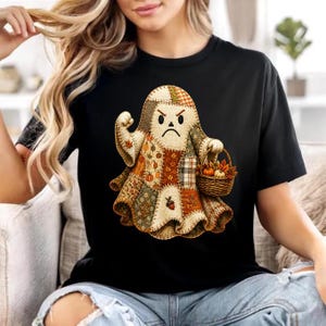 May include: A black t-shirt featuring a graphic of a ghost with a patchwork quilt pattern in shades of orange, brown, and cream. The ghost has an angry expression, one raised fist, and is holding a small woven basket filled with pumpkins.