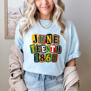 May include: Light blue t-shirt with the words "JUNE TEENTH 1865" in colorful block letters. The shirt is a crew neck style and is being worn by a person. The person is wearing a gold necklace and blue jeans.
