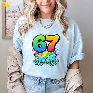 May include: Light blue t-shirt featuring a colorful graphic with the number "67" and two open hands. The number is in green, blue, and orange, with colorful paint splatters around the hands. A gold necklace is visible.