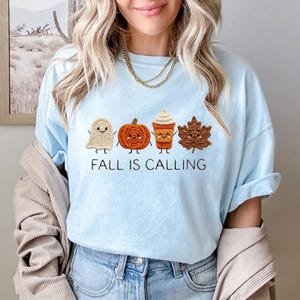 May include: A light blue t-shirt featuring a graphic of four crocheted-style characters: a white ghost, an orange pumpkin, a tan coffee cup, and a brown maple leaf. Below the characters, the text reads FALL IS CALLING in black capital letters.
