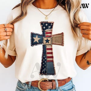 May include: A cream-coloured t-shirt with a patriotic cross design. The cross is made from denim with stars, red and white stripes, and burlap. The design is centrally placed on the shirt, creating a unique and eye-catching appearance.