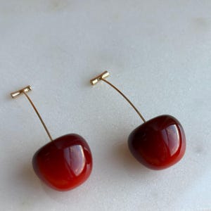 May include: A pair of cherry earrings. Each earring has a glossy, deep red cherry with a gold-coloured stem and post. The cherries are rounded and shiny, creating a playful and stylish accessory.