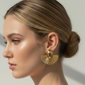 Pode incluir: Brincos dourados. Cada brinco apresenta uma parte superior oval e um design em forma de leque com linhas verticais. Os brincos são usados na orelha de uma pessoa, com o rosto e o cabelo da pessoa visíveis no fundo.