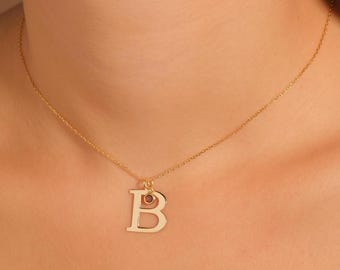 Könnte beinhalten: Eine goldfarbene Halskette mit einem Anhänger in Form des Buchstabens "B". Der Anhänger hat einen kleinen, runden, rotbraunen Stein. Die Halskette wird vor einem hautfarbenen Hintergrund gezeigt.