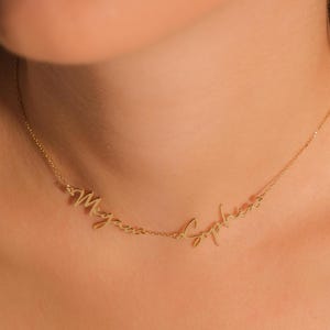 Op de afbeelding: Een delicate goudkleurige ketting met de namen "Megan" en "Sophia" in cursief schrift. De ketting wordt getoond tegen een huidkleurige achtergrond, wat het elegante ontwerp en het vakmanschap van de sieraden benadrukt.