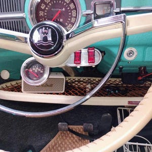 May include: The interior of a vintage vehicle featuring a turquoise dashboard, a cream-colored steering wheel with a central emblem, a circular speedometer, a fuel gauge, two red toggle switches, and a small rectangular speaker mounted below.
