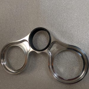 Puede incluir: Un componente metálico de color plateado con tres aberturas circulares, cuya abertura superior está revestida con un material negro texturizado. La superficie del metal muestra una pequeña letra R grabada en el lado derecho.
