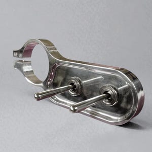 Puede incluir: Una placa de interruptores de metal pulido con dos interruptores de palanca. La placa tiene una abrazadera redondeada en un extremo y una base rectangular con los interruptores. Los interruptores tienen palancas plateadas.