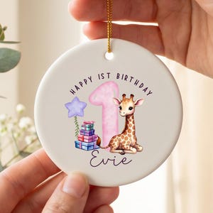 May include: A round white ceramic ornament featuring a watercolor illustration of a giraffe sitting next to a large pink number one. The design includes a purple star balloon, a stack of colorful gift boxes, and the text Happy 1st Birthday Evie.
