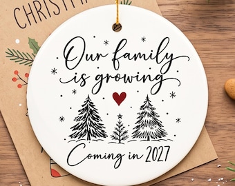May include: A round white ceramic ornament featuring black text that reads Our family is growing and Coming in 2027. The design includes three black pine tree illustrations, a small red heart, and snowflake accents, suspended by a gold string.