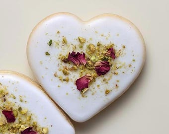 May include: Two heart-shaped cookies with smooth white icing, topped with a scattering of crushed green pistachio nuts and dried dark pink rose petals. The cookies have a light golden-brown edge and a rounded, symmetrical heart shape.