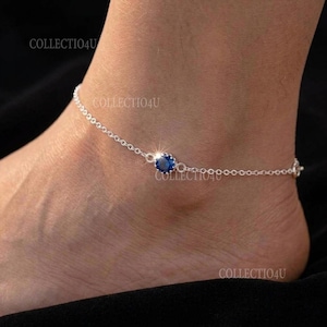 Op de afbeelding: Een zilverkleurig enkelbandje met een enkele ronde, gefacetteerde blauwe edelsteen in een klauwzetting. De fijne metalen schakels van de ketting verbinden zich met de centrale blauwe steen, wat zorgt voor een verfijnd sieraad voor de enkel.