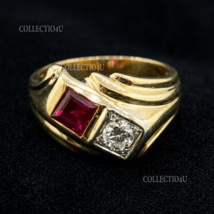 Puede incluir: Un anillo de oro con una gema roja cuadrada y un diamante cuadrado y transparente. El anillo tiene una banda gruesa con un diseño texturizado. El fondo es negro.