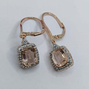 Puede incluir: Un par de pendientes de oro rosa con una piedra central cuadrada de color melocotón, rodeada por un halo de pequeñas piedras transparentes. Cierre de palanca.