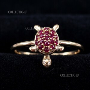 Puede incluir: Un anillo dorado con un diseño de tortuga. El caparazón de la tortuga está hecho de numerosas piedras preciosas pequeñas, redondas y de color rojo intenso. La cabeza y las aletas de la tortuga también son doradas, con dos pequeñas gemas blancas en la cabeza.