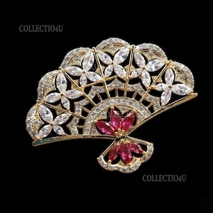 May include: A gold-tone fan-shaped brooch featuring clear marquise-cut stones arranged in floral patterns across the top and vibrant red marquise-cut stones at the base. The metal frame is accented with small clear stones. Text reads COLLECTIO4U.